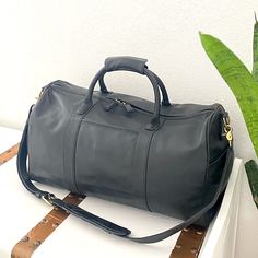 a black leather duffel bag sitting on top of a white table next to a plant