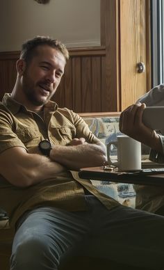 two men are sitting at a table with their arms crossed and one is holding a coffee mug