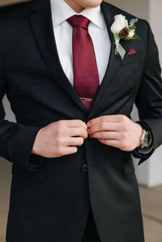 a man in a black suit and red tie is adjusting his lapel flower on his jacket