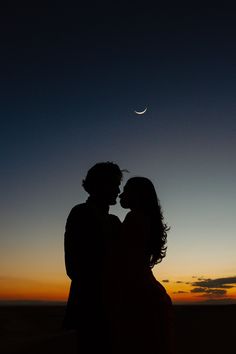 a man and woman standing next to each other in the desert at night with the moon behind them