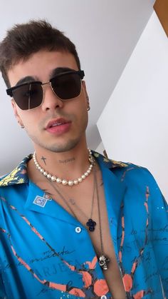 a young man wearing sunglasses and a blue shirt with flowers on the collar is standing in front of a white wall