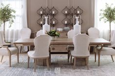 a dining room table with white chairs and vases on the wall above it, in front of two large windows