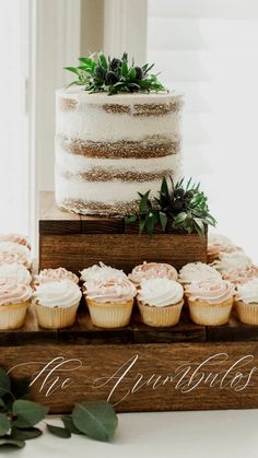 a wedding cake and cupcakes on a wooden stand with greenery in the background