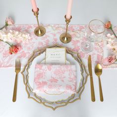 a place setting with pink flowers and gold cutlery on the table, along with candles