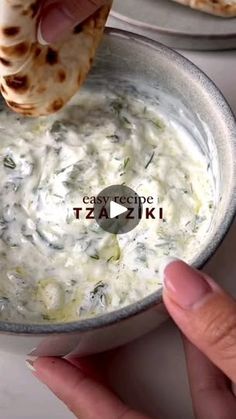 a person dipping some food into a bowl with pita bread on the side and another hand reaching for it