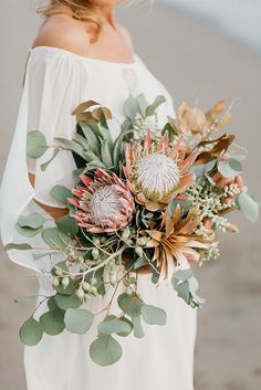 a woman holding a bouquet of flowers and greenery in her hands on the beach