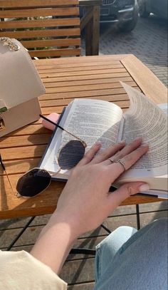 a woman is reading a book and sunglasses on a wooden table with her hand resting on an open book