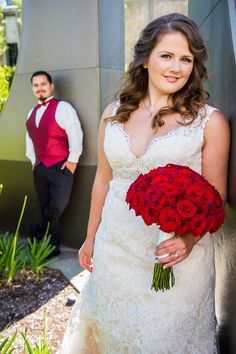 a bride and groom standing next to each other in front of a wall holding red roses