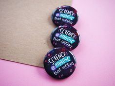 three badges with words on them sitting next to a piece of brown paper and pink background