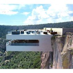 a house on top of a cliff in the middle of trees and mountains, with an outdoor dining area overlooking it