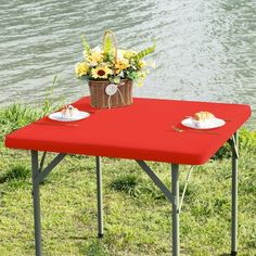 a red picnic table with flowers in a basket and bread on it next to the water