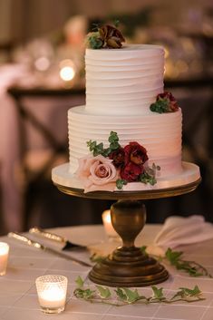 a white wedding cake with red flowers and greenery sits on a gold pedestal next to candles