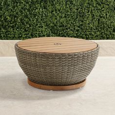 a round wooden table sitting on top of a cement floor next to a green hedge