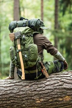 A person sitting on a large fallen tree trunk in a forest, viewed from behind. The person is wearing camouflage pants and a long-sleeve shirt, with a large green backpack. An axe is securely attached to the back of the backpack, and additional equipment, such as a knife in a sheath, is strapped to the person's belt. The forest background is lush and green, suggesting a serene and natural environment. The scene conveys a sense of preparedness and connection with nature. Acne Denim, Denim Projects, Star Wars Ships, Tactical Vest, Bicycle Design, Hiking Gear