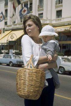 a woman walking down the street with a baby in her arms and a wicker basket over her shoulder