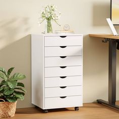 a white chest of drawers next to a potted plant on a wooden floor in front of a computer desk