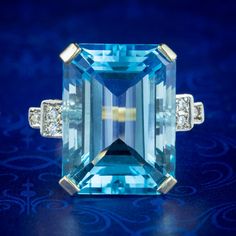 An extravagant Art Deco style cocktail ring boasting a mammoth emerald cut blue topaz, weighing an impressive 25 carats (approx.) It has a deep ocean blue hue, like an aquamarine and is flanked by three twinkling brilliant cut cubic zirconia set upon stepped shoulders. The topaz is held in a substantial four-claw mount that crowns it high and proud atop the finger. It's all silver and has been gilded in 18ct yellow gold. It's a fantastic statement piece and is sure to turn heads. WEIGHT: 10 gram Luxury Art Deco Topaz Rings, Classic Blue Octagon Topaz Ring, Art Deco Blue Topaz Ring For Anniversary, Blue Art Deco Topaz Ring For Anniversary, Luxury Turquoise Topaz Ring For Formal Occasions, Rectangular Blue Topaz Ring With Diamond Accents, Luxury Blue Baguette Cut Topaz Ring, Luxury Formal Turquoise Topaz Ring, Blue Octagon Art Deco Jewelry