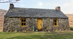 an old stone house with yellow doors and windows on the side of a grassy hill