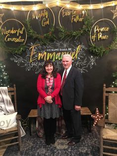 two people standing next to each other in front of a christmas sign with evergreen decorations