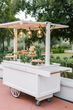 a white cart with food on it and lights hanging from the roof, in front of trees