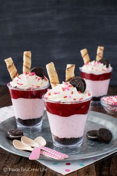 three dessert cups with oreo cookies on top and red liquid in the middle, topped with whipped cream and sprinkles