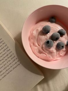 a pink bowl filled with blueberries next to an open book