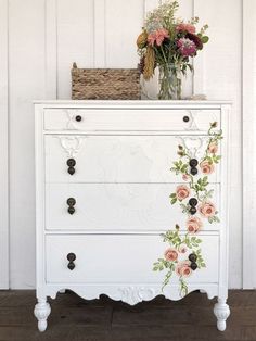 a white dresser with flowers painted on the top and bottom drawers, next to a basket full of flowers