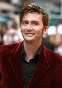 a man in a red velvet jacket and black shirt smiles at the camera as people look on