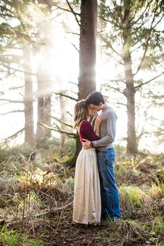 a man and woman standing in the woods hugging each other with sunlight shining through the trees behind them