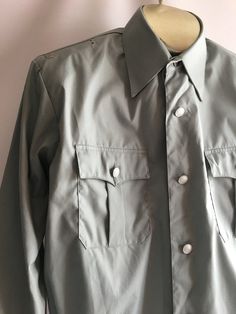 Awesome vintage men's uniform work shirt jacket. The military inspired cut and color encourage me to believe this was a aviation uniform of some sort. Somewhere between a shirt and a jacket, these piece has some great details. The material is most likely a permanent press style poly/cotton blend, though no content label is present. The shirt has a button front styling much like a classic men's dress shirt but with a more boxy cut and elastic waist. It has small loops and holes near top shoulder Military Style Button-up Work Shirt, Military Style Button-up Shirt For Work, Military Style Shirt For Work With Button Closure, Military Style Workwear Button-up Shirt, Long Sleeve Workwear Uniforms, Military Long Sleeve Work Shirt, Military Style Shirt With Buttons For Work, Military Style Collared Shirt For Workwear, Military Style Workwear Shirt With Buttons