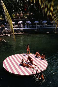 several people are in the water on a pink and white striped floating platform that is surrounded by palm trees