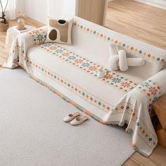 a white couch covered with a blanket and pillows on top of wooden flooring next to a window