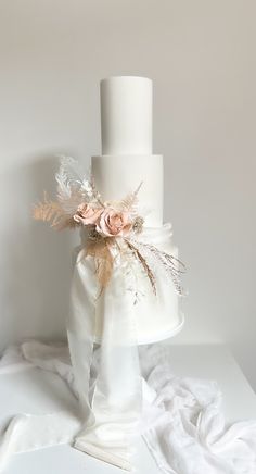 a three tiered white wedding cake with pink flowers and feathers on top, sitting on a table