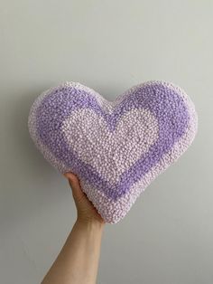 a hand holding a purple and white knitted heart shaped pillow on top of a gray wall