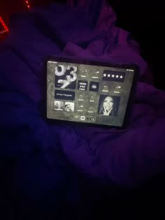 a tablet sitting on top of a bed covered in purple sheets and blankets, with the screen lit up