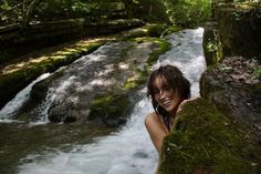 a woman standing next to a river in the forest with green mossy rocks and trees