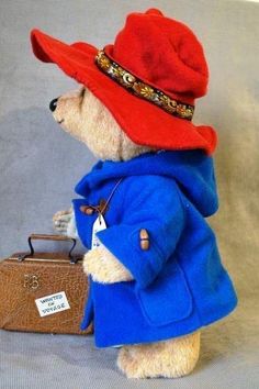 a teddy bear wearing a red hat and blue coat with a suitcase on the ground