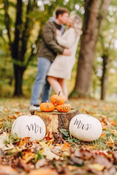 an engaged couple standing next to pumpkins on the ground in front of some trees