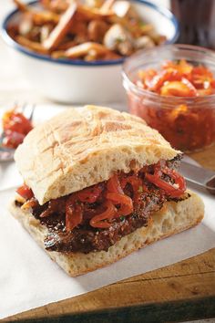 a sandwich with meat, tomatoes and onions on a wooden cutting board next to a bowl of cole slaw