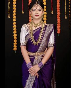 a woman in a purple and gold sari with beads around her neck, standing next to an orange garland