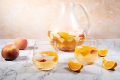 two glasses filled with liquid next to sliced oranges and apples on a marble table