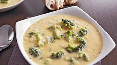 two bowls of broccoli and cheese soup on a table with spoons next to it