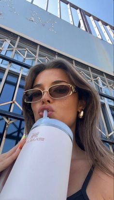 a woman wearing sunglasses drinking from a water bottle while standing in front of a building