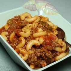 a white bowl filled with pasta and meat on top of a blue table cloth next to a spoon