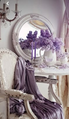 a white table topped with purple flowers next to a chair and window covered in curtains