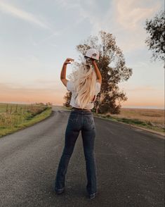 a woman in jeans and a hat standing on the side of a road with her back to the camera