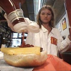 a woman pouring ketchup onto a hot dog bun on top of a table