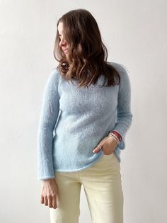 a woman standing in front of a white wall wearing a blue sweater and cream pants