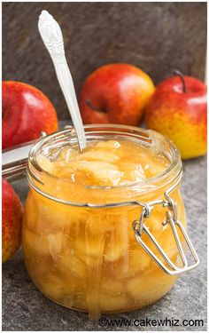 an apple jam in a glass jar with two spoons next to it and apples