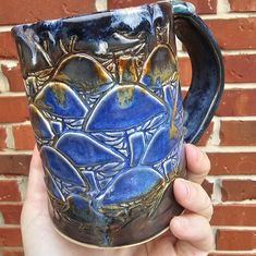 a hand holding up a blue and black coffee mug with designs on the outside, in front of a brick wall